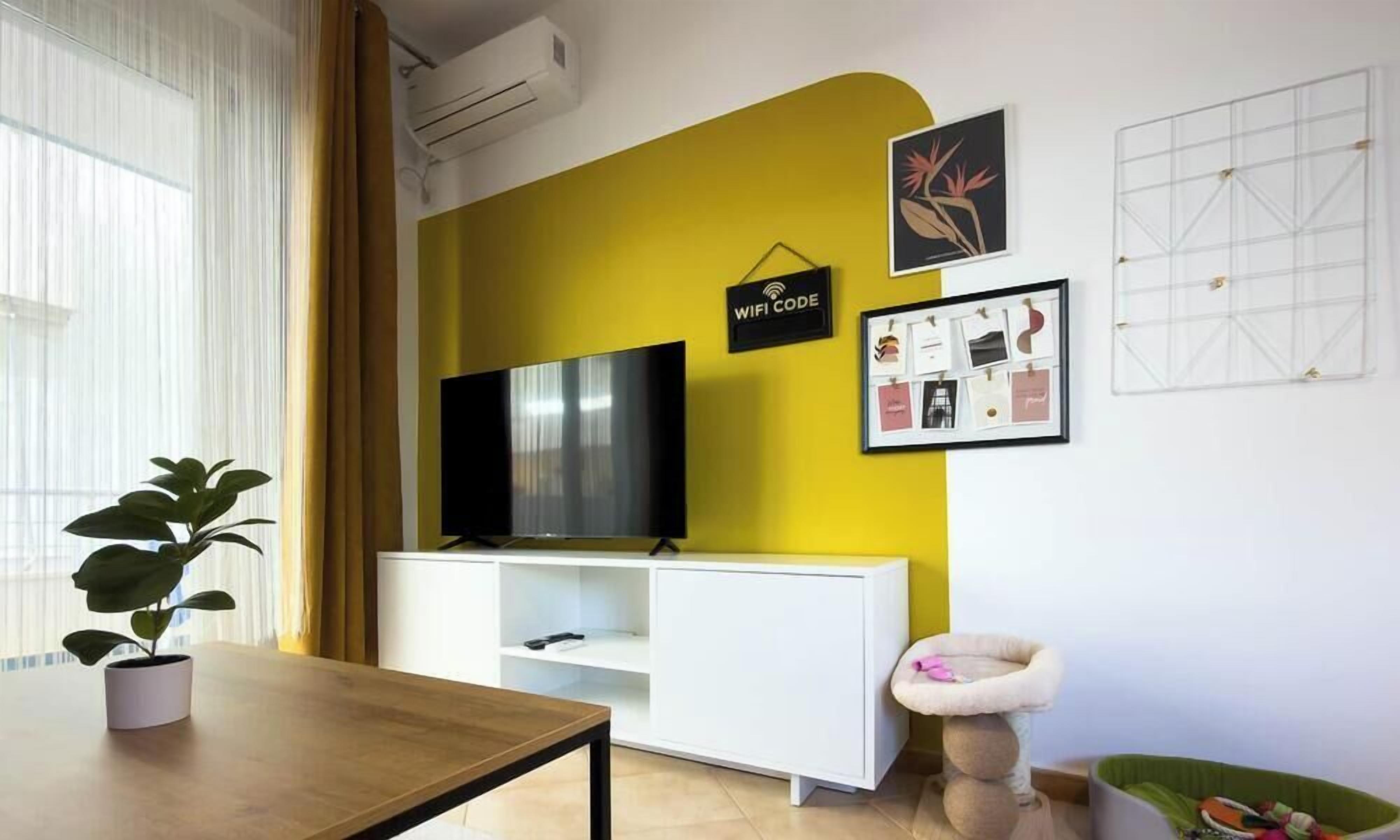 Un salon moderne avec un mur d’accent jaune, un téléviseur à écran plat posé sur un meuble blanc, une plante en pot, un panier pour chat et un ensemble de photos encadrées.