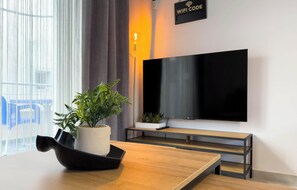A modern room with a wooden table, a potted plant, a flat-screen TV mounted on the wall, and a sign indicating a WiFi code. - Amelia Beach (Pet Friendly) (Sarandë)