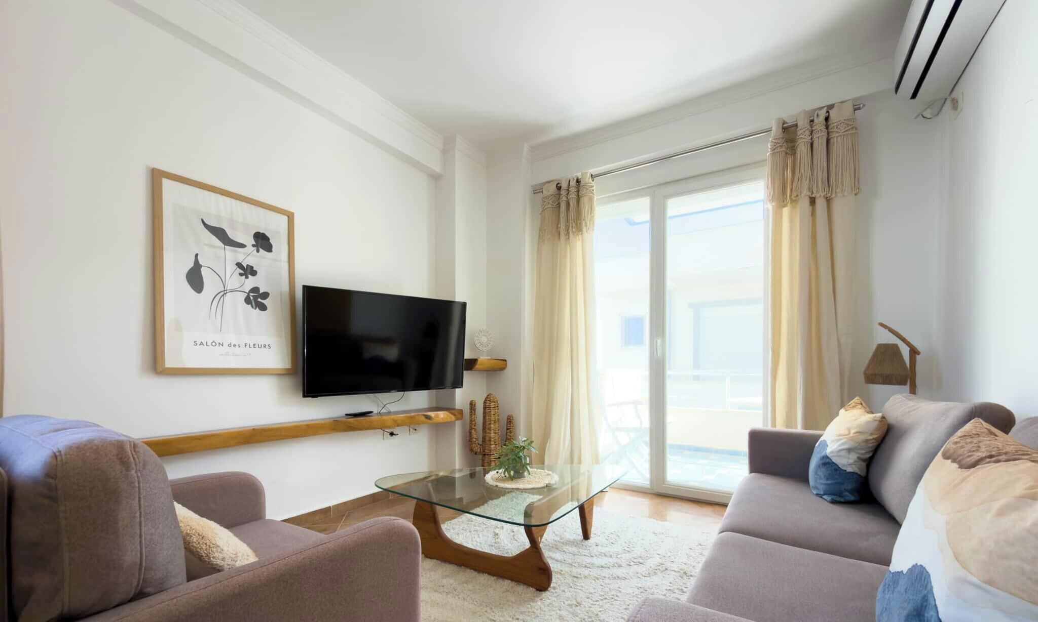 A modern living room with a flat-screen TV, two sofas, a glass coffee table, and a large window with curtains.