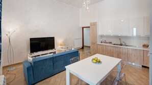 Ein modernes Wohnzimmer mit Flachbildfernseher, einem blauen Sofa, einem Esstisch mit Obstschale und einer Küchenzeile mit Holzschränken und einer weißen Arbeitsplatte.