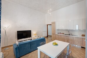 Un soggiorno moderno con una televisione a schermo piatto, un divano blu, un tavolo da pranzo con una ciotola di frutta e un angolo cottura con mobili in legno e un piano di lavoro bianco.