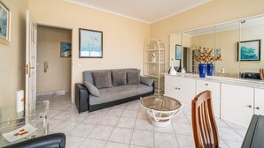 Ein Wohnzimmer mit einer Couch, einem Glastisch, einem Esstisch mit Stühlen, einem weißen Schrank und einem großen Spiegel.