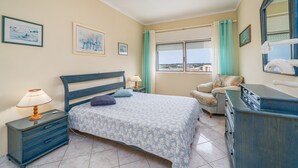 Ein Schlafzimmer mit Bett, Nachttisch, Schminktisch, Stuhl, einem Fenster mit Vorhängen und Kunstwerken an den Wänden.