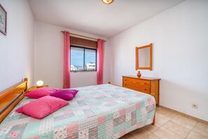 Un dormitorio con cama, cómoda, ventana con cortinas, lámpara de noche y espejo montado en la pared.