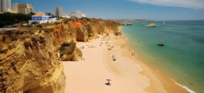 Una playa con un acantilado rocoso, agua cristalina y edificios al fondo.