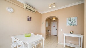 Uma área de refeições com uma mesa e cadeiras brancas, um relógio de parede e uma pequena mesa com um objeto decorativo.