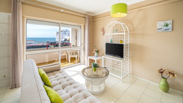 Uma sala de estar com sofá, uma mesa de vidro redonda, uma TV sobre um suporte branco, um vaso verde com flores e vista para o mar através de uma janela grande.