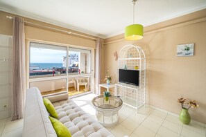 Un saló amb un sofà, una taula rodona de vidre, una televisió sobre un moble blanc, un vas verd amb flors i vista al mar a través d'una finestra gran.