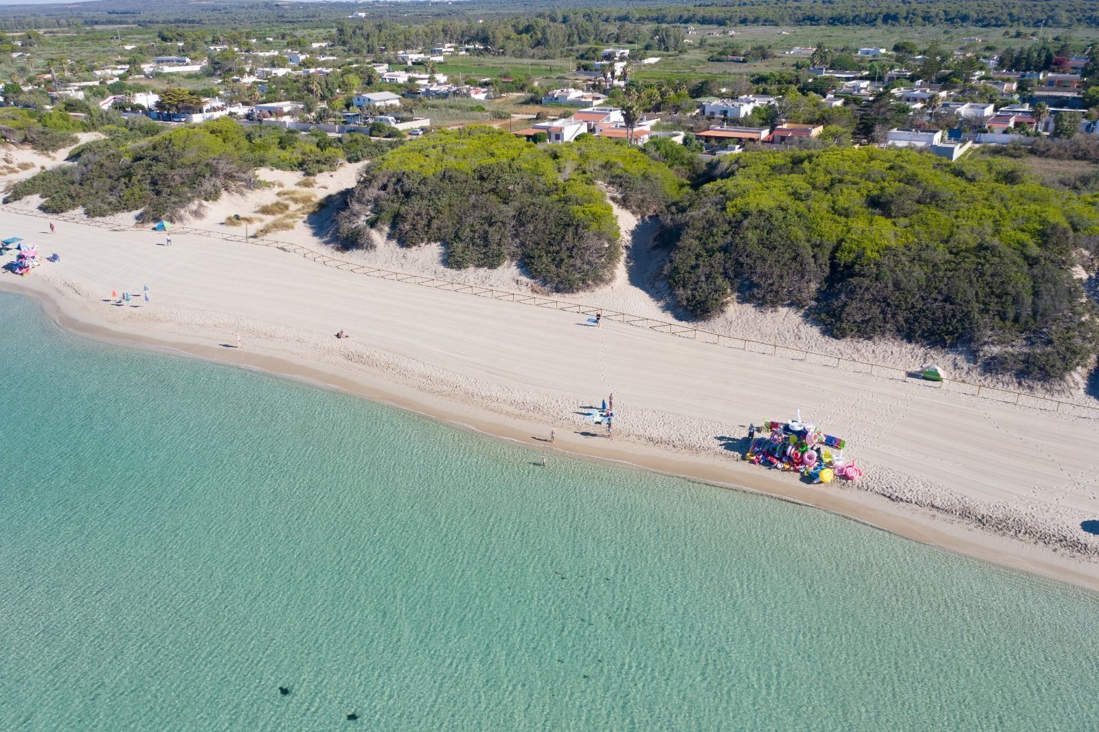 Vue aérienne d’une zone côtière avec une plage de sable, une eau turquoise et limpide, et une zone résidentielle avec des maisons et de la végétation en arrière-plan.