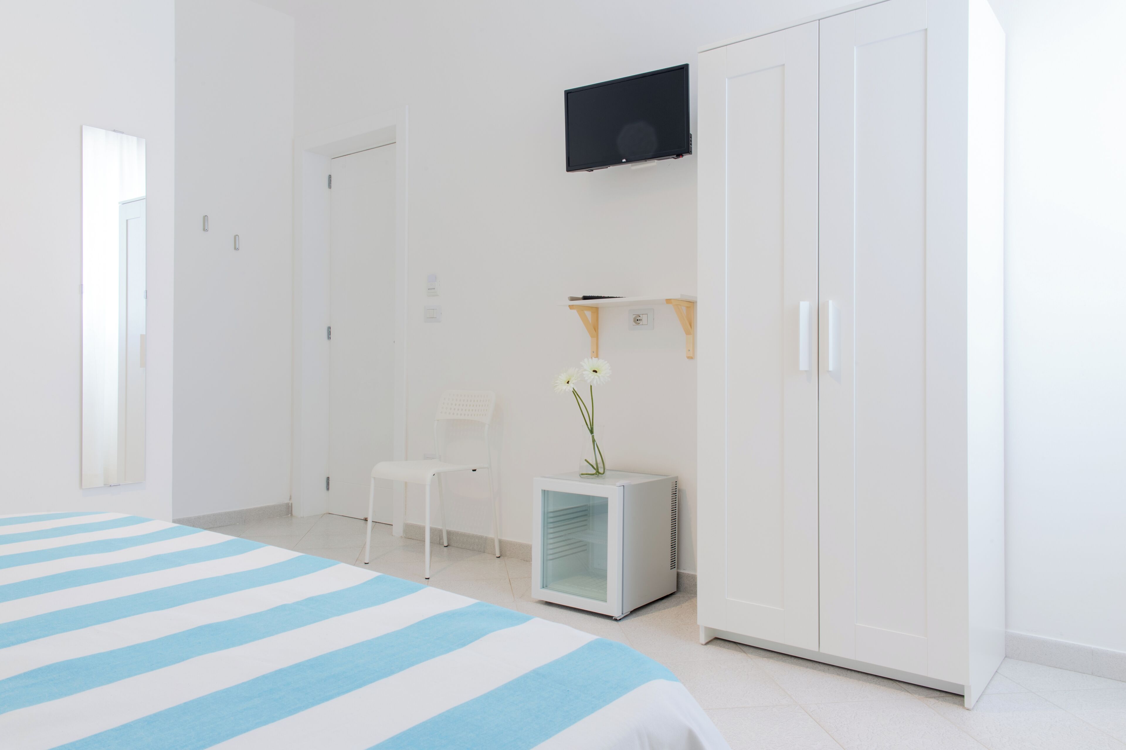 A hotel room with a bed, a TV, a wardrobe, a chair, and a small table with a flower.