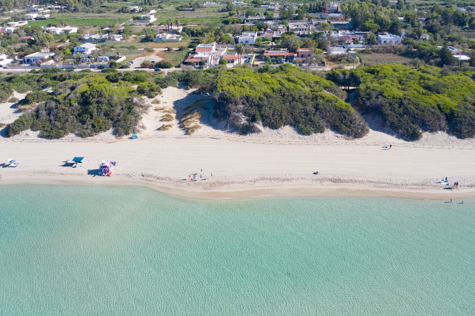 Vue aérienne d’une zone côtière avec une plage de sable, des eaux turquoise et limpides, et une zone résidentielle avec des maisons et de la végétation en arrière-plan.