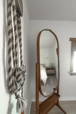 Un dormitorio con un espejo de madera, una cama con ropa de cama blanca, una mesita de noche con una lámpara y un tapiz colgado en la pared.