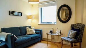 A living room with a blue sofa, a plaid armchair, a small table with a lamp, a mirror, and a window with blinds.