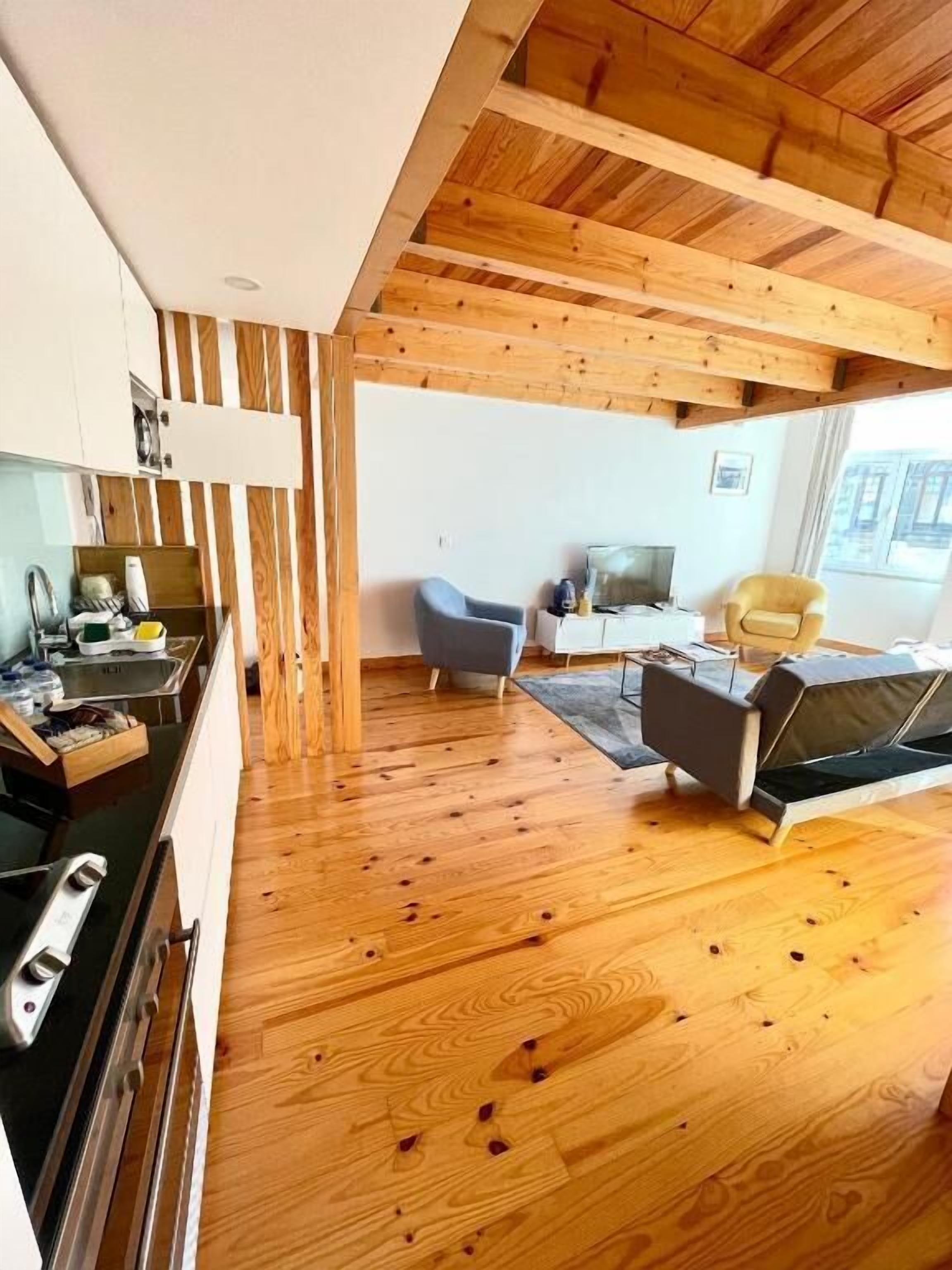 Una cocina moderna con vigas de madera, una zona de comedor con sofá y televisor, y un amplio ventanal que proporciona luz natural.