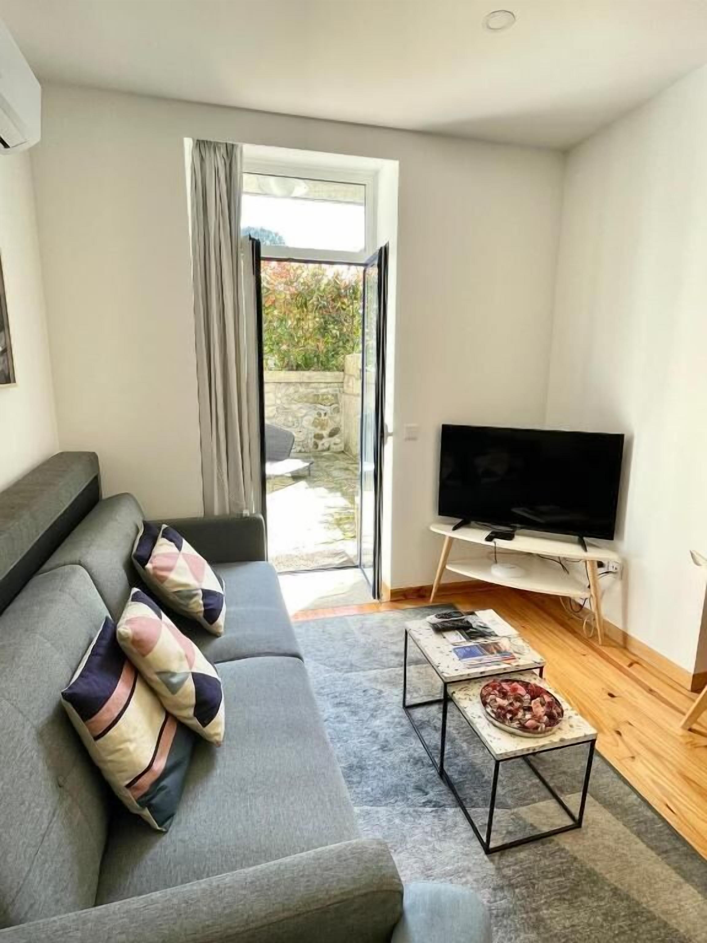 A living room with a grey sofa, a flat-screen TV on a wooden stand, and a small coffee table with magazines.