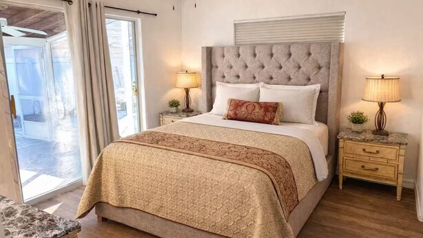 Une chambre à coucher avec un grand lit, deux tables de chevet avec des lampes, une porte-fenêtre coulissante et un ventilateur de plafond.