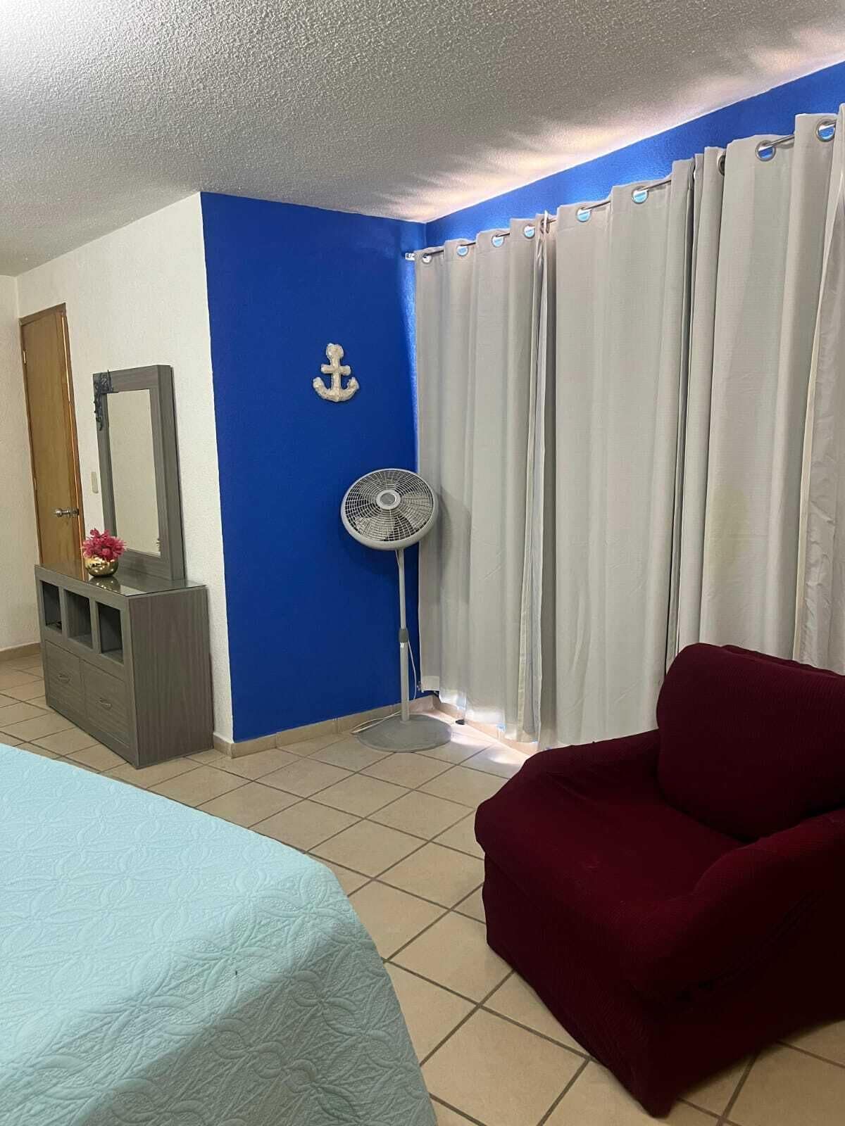 A room with a bed, a red chair, a fan, a mirror, and curtains.