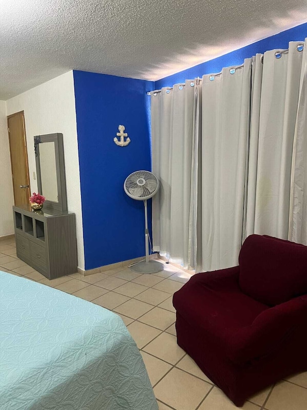 Une pièce avec un lit, une chaise rouge, un ventilateur, un miroir et des rideaux.