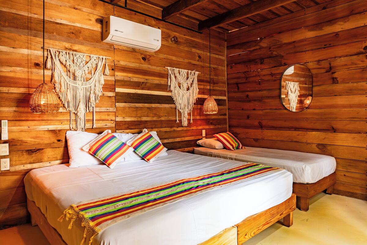 A wooden cabin-style room with a large bed, two smaller beds, macramé wall hangings, and a round mirror.