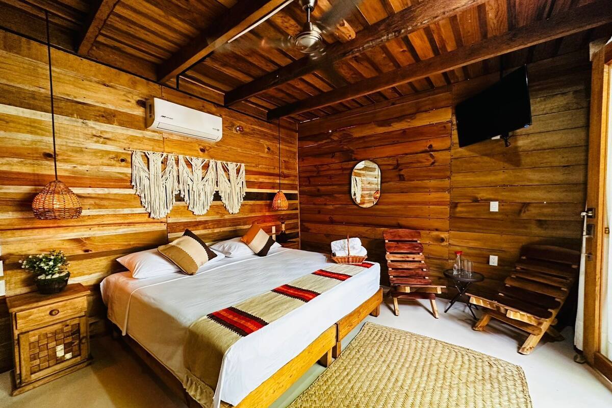A wooden cabin-style bedroom with a bed, a small table, a chair, a woven rug, and a ceiling fan.