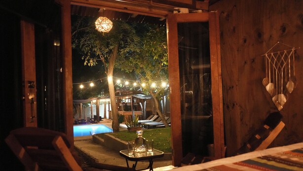 Une porte ouverte donne sur un espace piscine agrémenté de guirlandes lumineuses, une table avec un plateau en verre et un hamac.