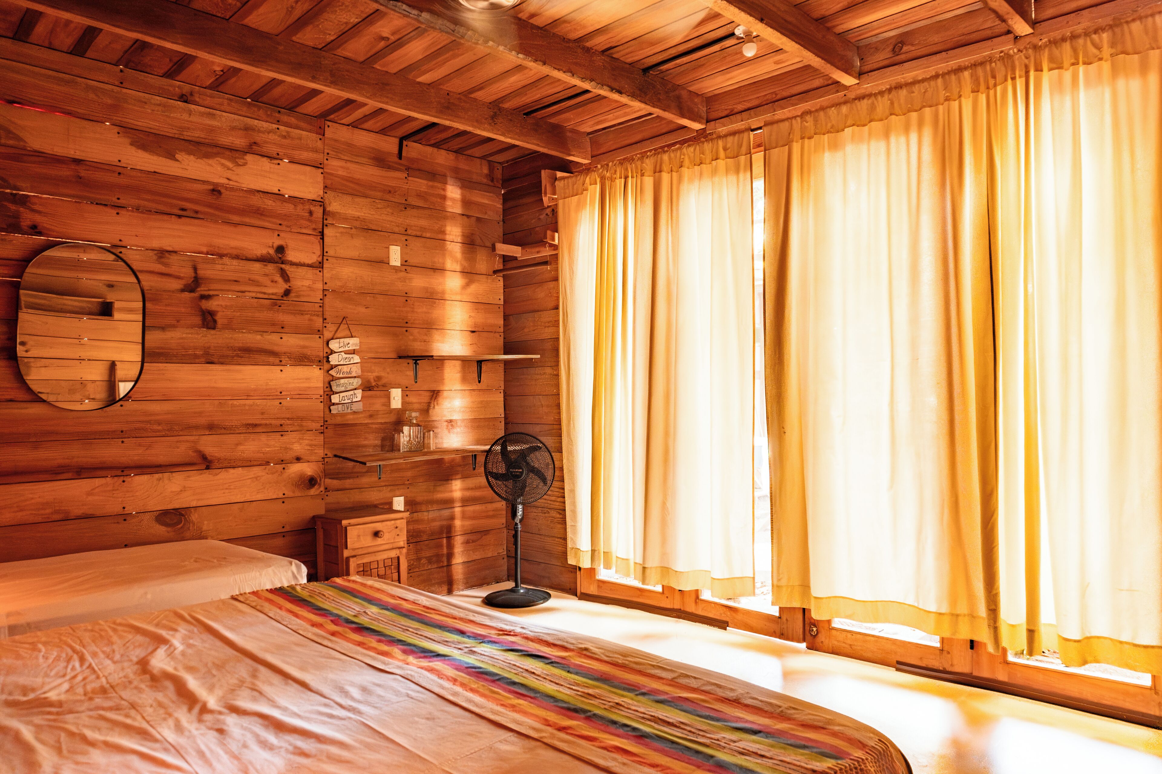 A wooden cabin room with a bed, a fan, a mirror, and curtains.