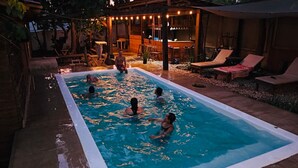 Une piscine rectangulaire où des personnes nagent, entourée de chaises longues en bois et d’un espace bar.