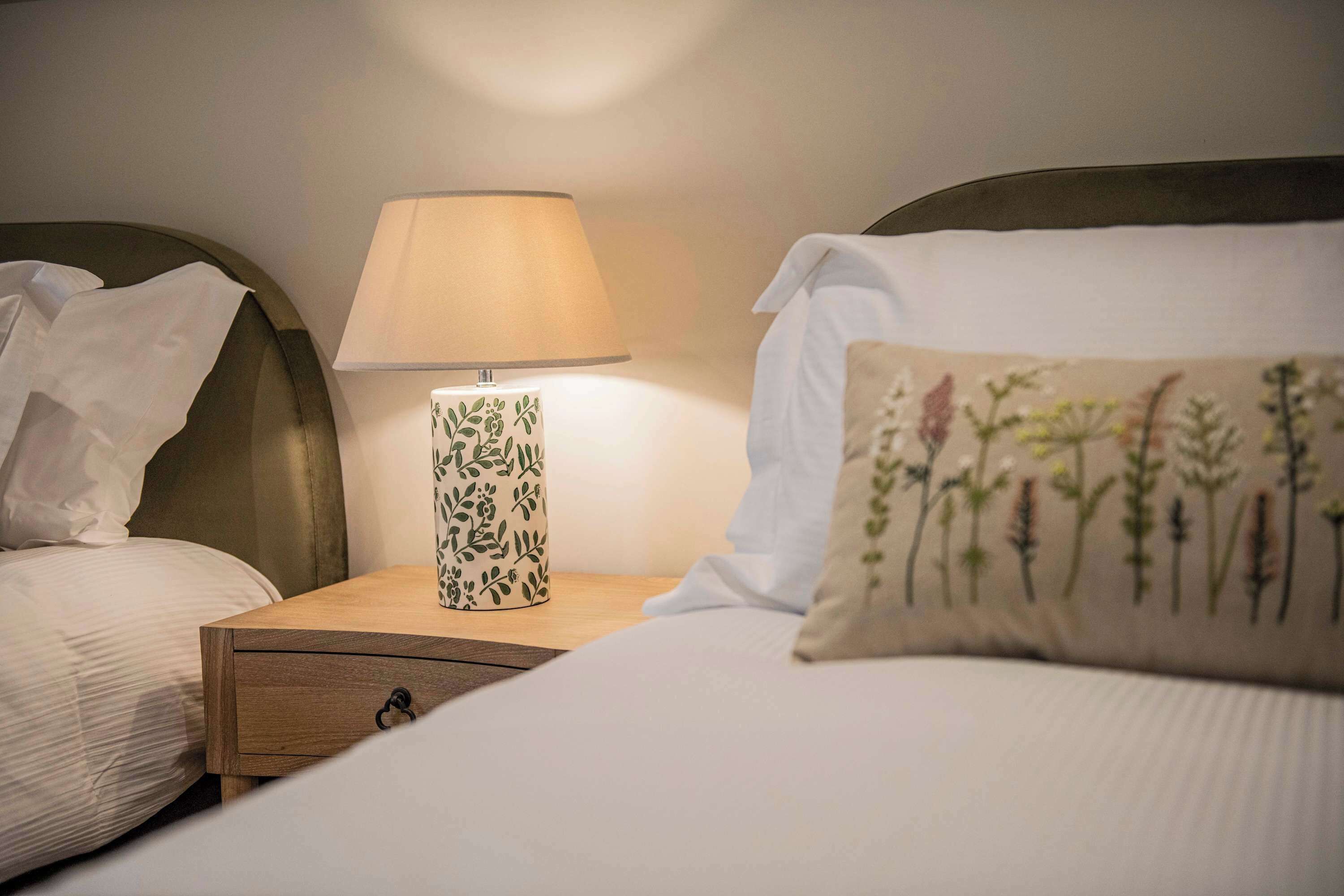 Een net opgemaakt bed met witte lakens en een sierkussen met bloemen, een bedlamp met een bladmotief, en een houten nachtkastje met een lade.