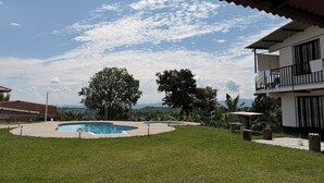 Une piscine entourée d’une terrasse, un bâtiment à deux étages doté de balcons et une vue sur des collines lointaines.
