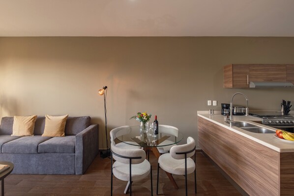 A modern living room with a sofa, a glass dining table, and a kitchen area with a stove and sink.