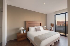 Una habitación de hotel moderna con una cama grande, cabecera de madera, dos mesitas de noche con lámparas y vistas a un paisaje urbano a través de un ventanal.