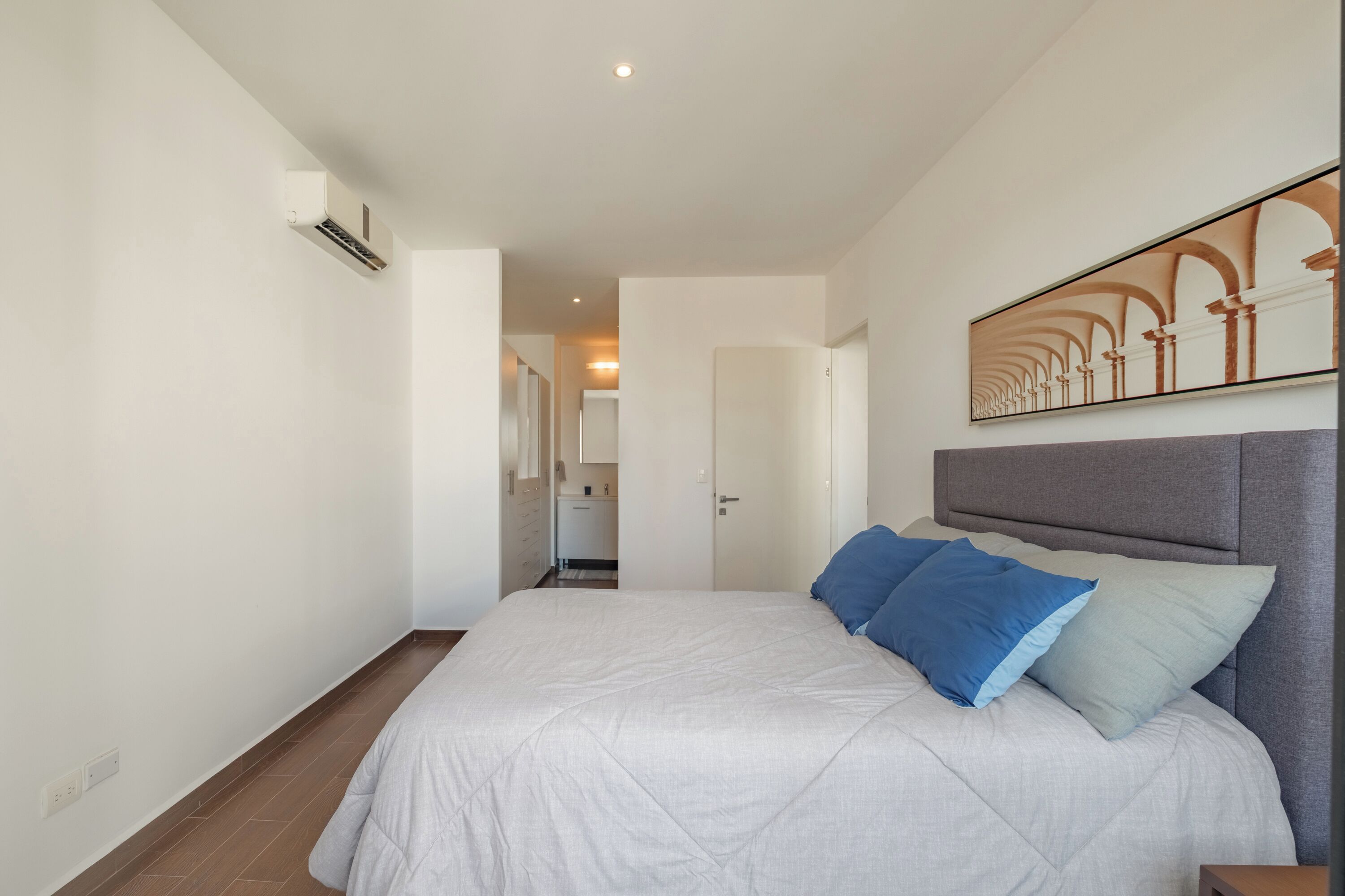 A bedroom with a bed, a headboard, a wall-mounted artwork, and a door leading to a bathroom.
