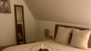 Una cama bien hecha con un arreglo de pétalos de rosa y velas, una mesita de noche con una lámpara, y un espejo en la pared.