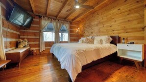 A log cabin bedroom with a wooden ceiling, a bed with white bedding, a bedside table, a nightstand, a TV mounted on the wall, and a window with curtains. - Near Toccoa River • Hot Tub • Fire Pit • Gameroom - Holiday Home (Blue Ridge)