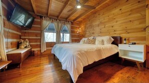 A log cabin bedroom with a wooden ceiling, a bed with white bedding, a bedside table, a nightstand, a TV mounted on the wall, and a window with curtains. - Near Toccoa River • Hot Tub • Fire Pit • Gameroom - Holiday Home (Blue Ridge)