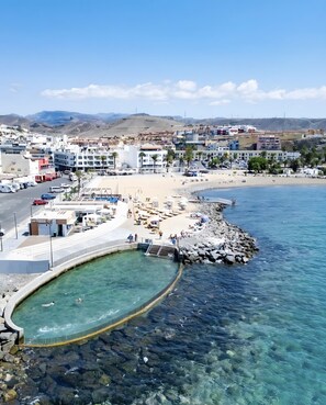 A coastal town with a clear blue sea, a rocky pier, and a sandy beach. - Sunset Star Holiday Home: Sea Views from Rooftop Solarium, Shared Terrace & Wi-Fi (Arguineguín)