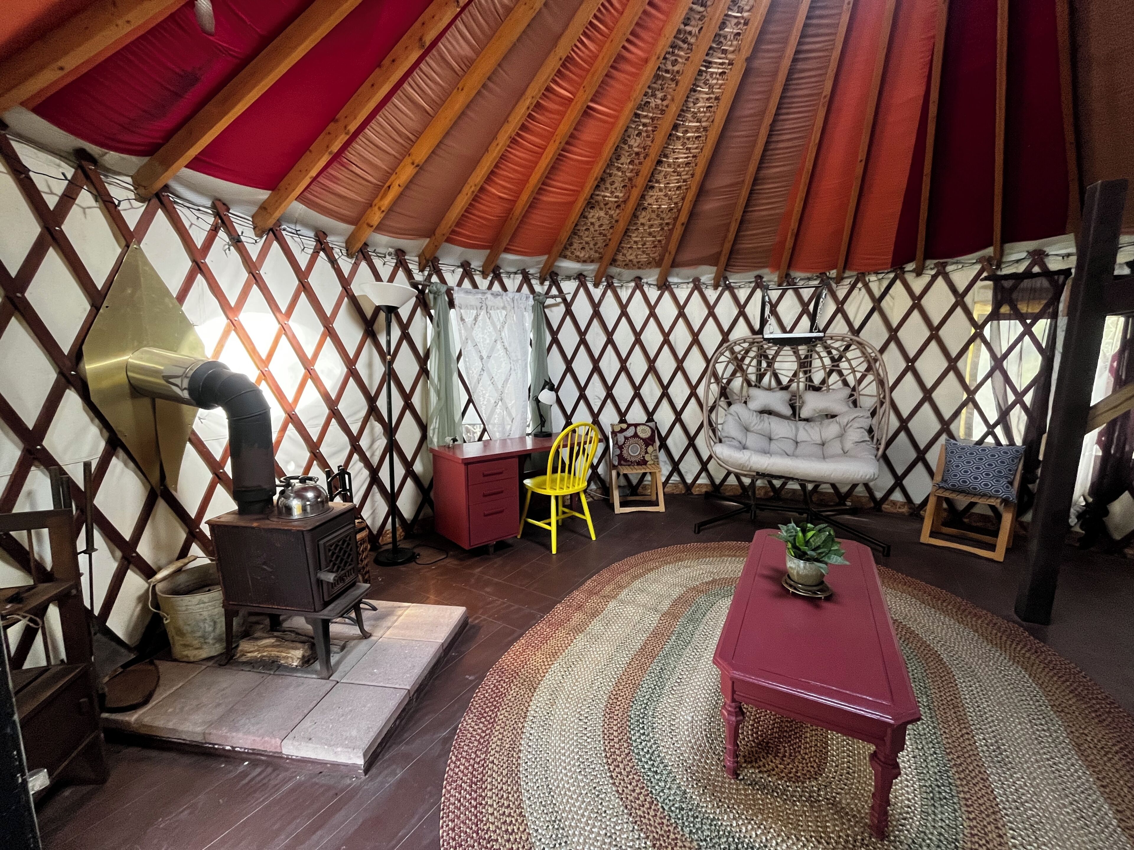 Interior de una yurta con un horno, una mesa roja, una silla amarilla y un sillón colgante.