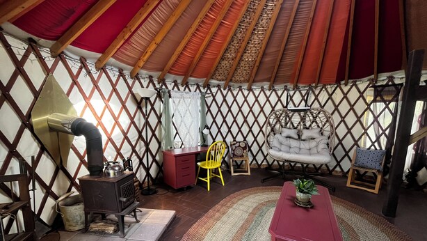 A yurt interior with a stove, a red table, a yellow chair, and a hanging chair. - Relax in a Cozy, Skylit Yurt at a Farm Animal Sanctuary (Pittsboro)