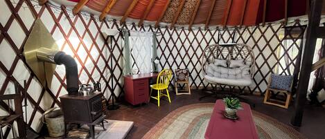 Interior de una yurta con un horno, una mesa roja, una silla amarilla y un sillón colgante.