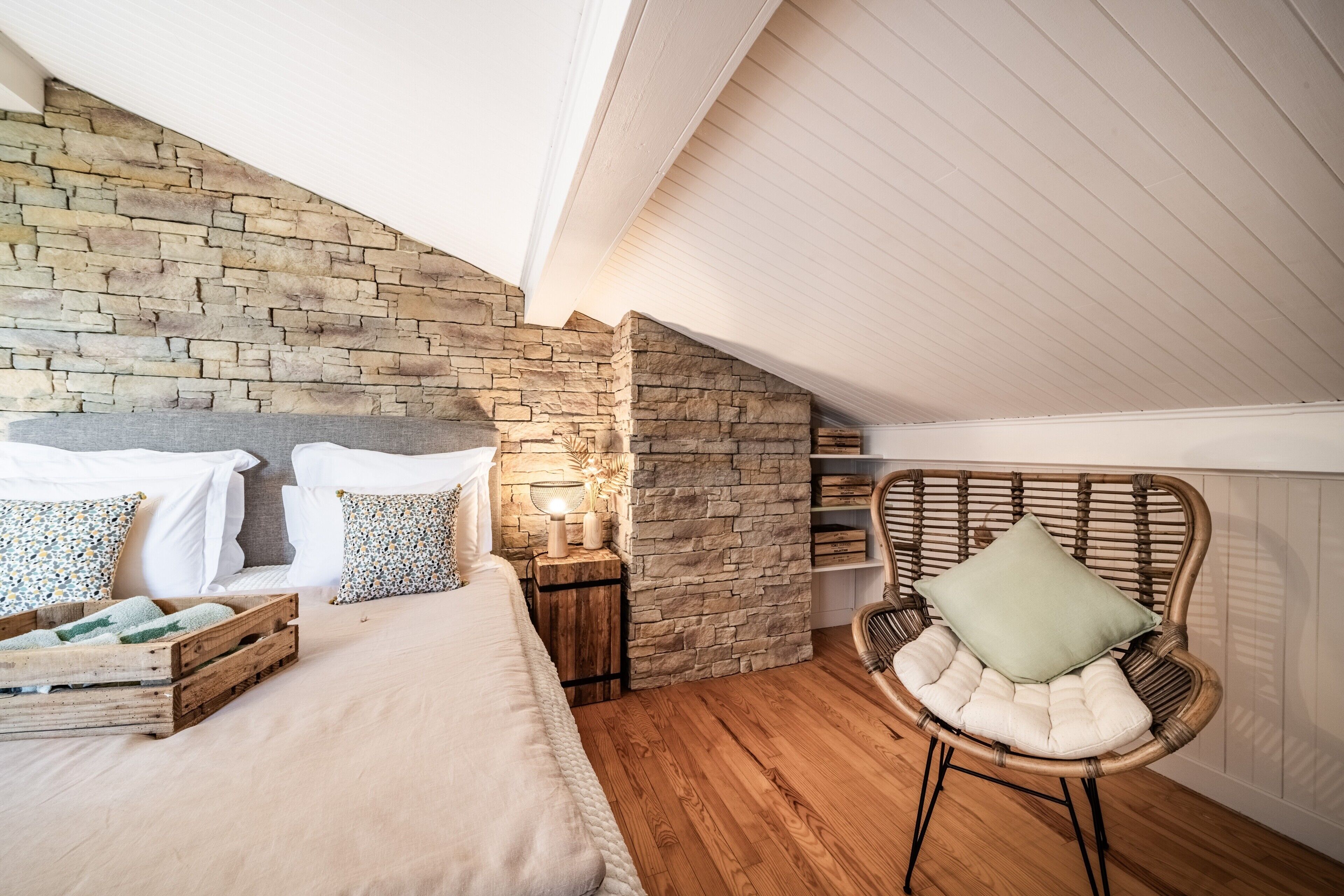 Ein Schlafzimmer mit Steinwand, Holzboden, einem Bett mit weißer Bettwäsche, einem Korbstuhl und einem Nachttisch mit Lampe.