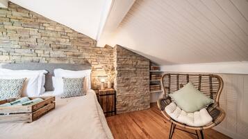 Ein Schlafzimmer mit Steinwand, Holzboden, einem Bett mit weißer Bettwäsche, einem Korbstuhl und einem Nachttisch mit Lampe.