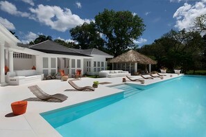 Ein moderner Poolbereich im Freien mit Liegestühlen, einer Struktur mit Reetdach und einem weißen Gebäude mit mehreren Sitzbereichen.