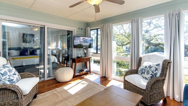 A spacious living room with wicker furniture, a ceiling fan, a television, and large windows offering a view of the outdoors. - 764 Summerwind Villa | Peaceful Edisto Escape | Golf, Lagoon & Wildlife Views | Easy Beach Access (Edisto Beach)