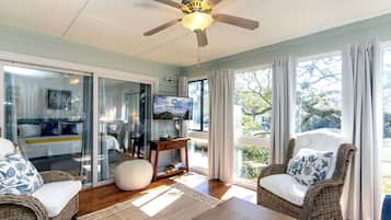 A spacious living room with wicker furniture, a ceiling fan, a television, and large windows offering a view of the outdoors.