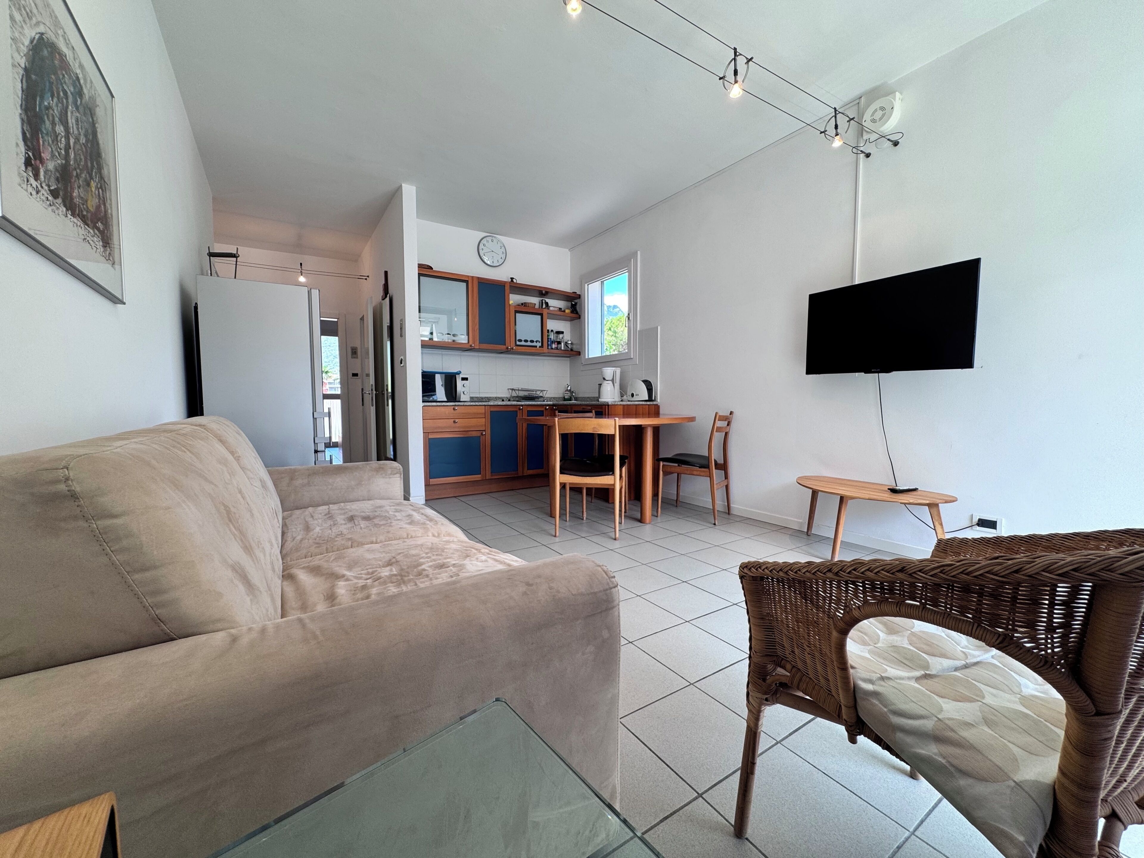 A living room with a sofa, a wicker chair, a television mounted on the wall, and a kitchen area in the background.