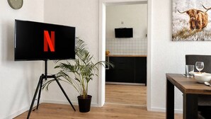 A modern living room with a large flat-screen TV displaying the Netflix logo, a potted plant, a wooden dining table with a bowl and glasses, and a kitchen visible through an open doorway. - N&N: Familienwohnung im Zentrum + Spielzeug (Schwerin)