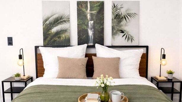 Un lit bien fait, recouvert d’une couverture verte, avec deux oreillers, un plateau sur lequel se trouvent un vase et des tasses, et trois tableaux encadrés accrochés au mur.