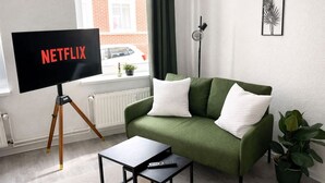 Un salon avec un canapé vert, des coussins blancs, une table basse noire et un téléviseur posé sur un trépied.