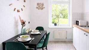Une cuisine moderne avec une table à manger pour deux personnes, une fenêtre donnant sur un paysage verdoyant et une horloge murale dont les chiffres sont en bois.