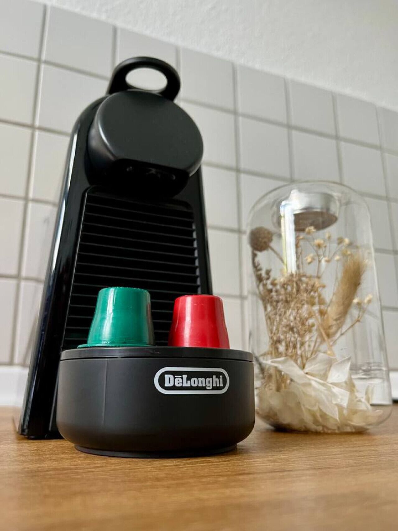 En svart vattenkokare från De'Longhi med ett genomskinligt lock av glas, innehållande torkade blommor, placerad på en träyta mot en vägg täckt med kakel.